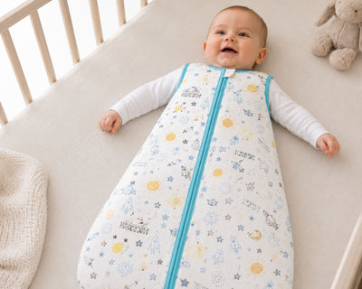A baby lying on a crib mattress wearing a sleep sack with a blue zipper and a white background printed with yellow and blue stars, moons, and suns. A teddy bear is visible in the corner of the crib.